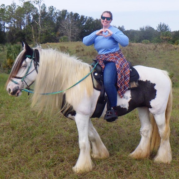 Ashley horseback riding on a trail, holding up a heart symbol