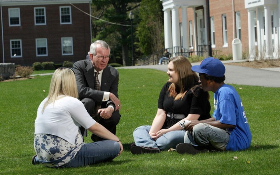 Three students sitting on a school lawn talking with a man dressed in professional attire